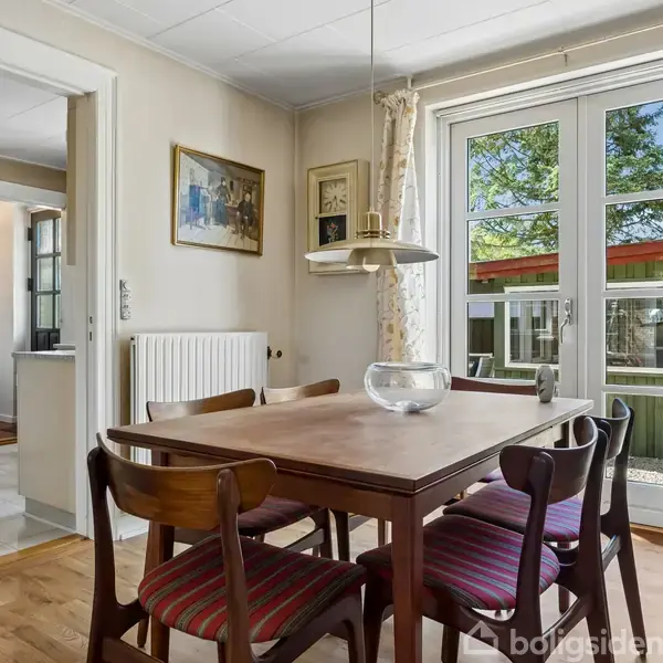 Et spisebord med seks stole står i et lyst rum. En lampe hænger over bordet. Gennem glasdøren ses en terrasse og have. Malerier hænger på væggen.