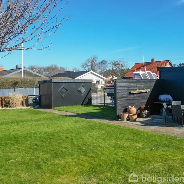 Et træ står stille i en grøn have ved siden af en terrasse med havemøbler og lerkrukker. I baggrunden ses flere huse under en blå himmel.