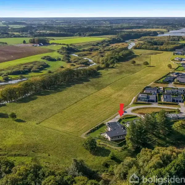 Et hus markeret med en rød pil ligger i et åbent, grønt landskab ved siden af en række andre huse. Markerne strækker sig i baggrunden under en klar blå himmel.