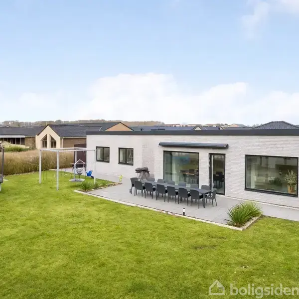En moderne villa med fladt tag står stille i en anlagt have. En stor terrasse har et langbord med stole, og der ses en trampolin og klatrestativ i græsplænen.
