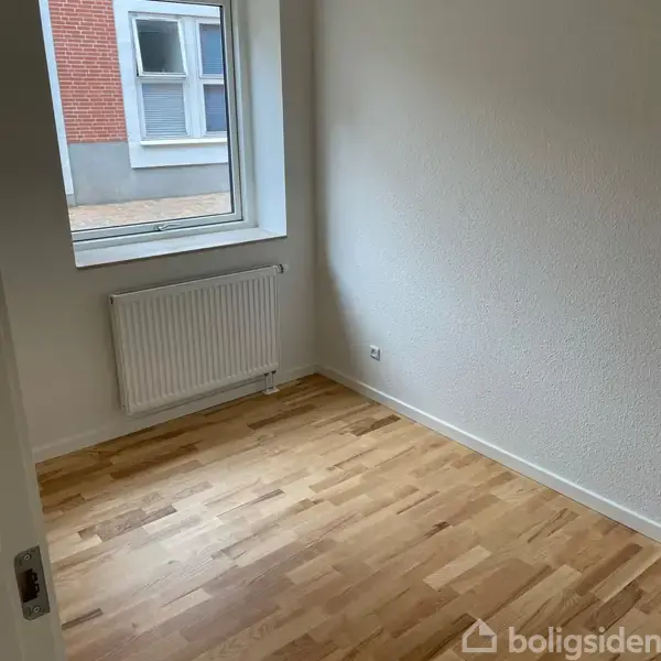 Et tomt rum med et trægulv og en radiator under et vindue. Udendørs ses en murstensvæg og en flisebelagt gangsti. Væggene er hvide, og der er intet inventar.