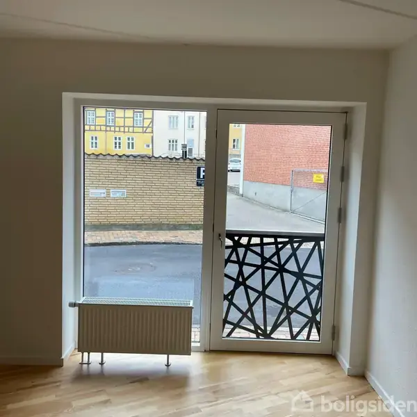 En glasdør med sort mønster fungerer som altandør. Udsigt til gaden med fortov, en murstensvæg samt boliger i baggrunden. Skiltet viser "P" parkeringssymbol.