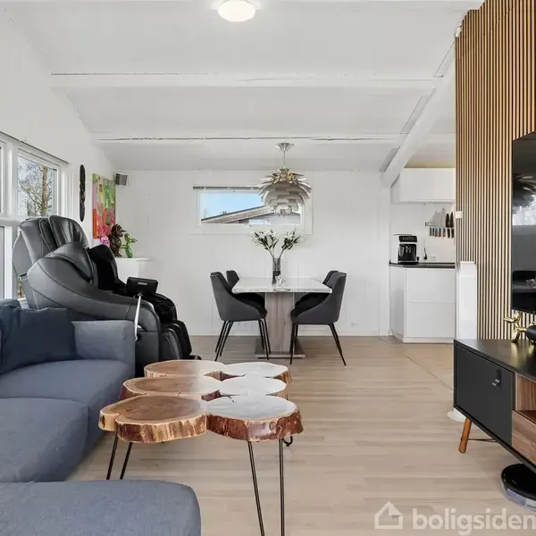Grål sofa står i stue med hvide vægge, træbord med naturkant foran. Sort lænestol ved vindue. Spisebord med fire stole i baggrund, moderne indretning. Ingen tekst.
