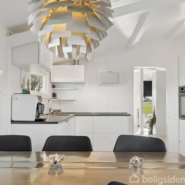 Et moderne køkken med hvide skabe og sort bordplade. En stor, dekorativ lampe hænger over et spisebord med sorte stole. I venstre hjørne står en vase med hvide blomster.