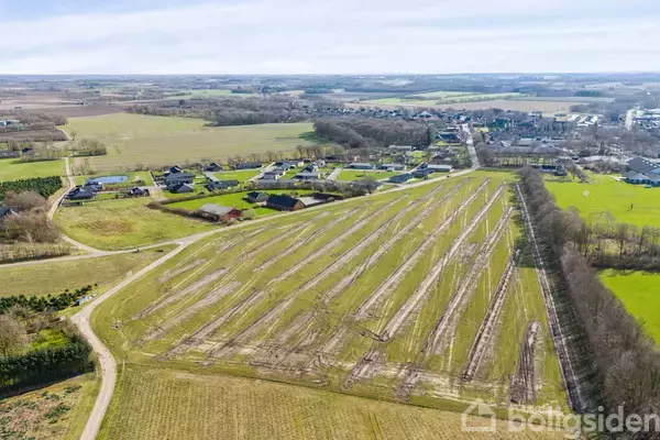 En stor mark med parallelle spor strækker sig gennem et åbent landskab. Omgivende huse og træer ses i baggrunden under en klar himmel.