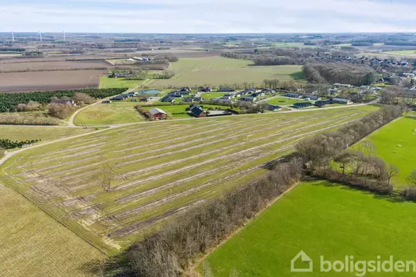 En stor mark ses fra luften med rækker af afgrøder. Baggrunden består af en landsby med huse og spredte skovområder under en åben, lys himmel.