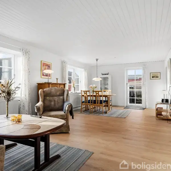 En lys stue med trægulv, hvor en lænestol og bord står ved vinduer. Et spisebord med stole ses i baggrunden, omgivet af store vinduer og en dør til terrassen.