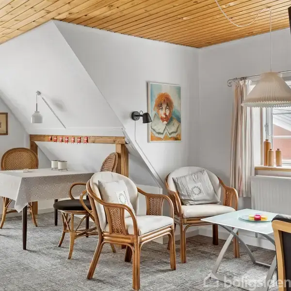 Rattanmøbler står i en stue med et hvidt bord, mens en vægmaleri og loftslamper skaber hygge. Der er en spisestue bagved med et dækket bord og stole.