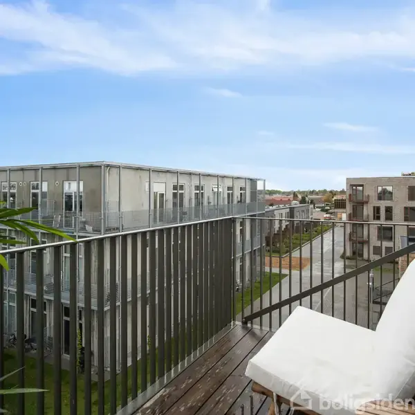 En balkon med en blød hvid pude ligger stille med udsigt over moderne lejlighedsbygninger og en klar himmel i en bynær omgivelser.