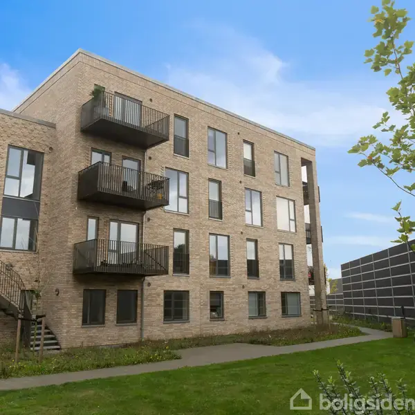 En moderne murstensbygning med altaner står i en grøn have. Træer og en stensætning omkranser bygningen under en klar blå himmel.