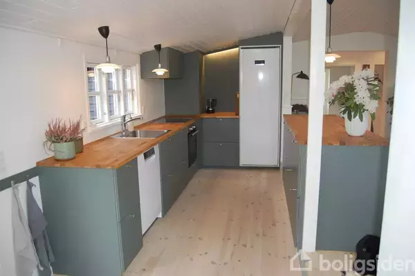 Moderne køkken med grå skabe, træbordplader og indbyggede apparater. Køkkenet er lyst op med pendellamper og har træplankegulv. En vase og blomsterdekoration pryder bordpladen.