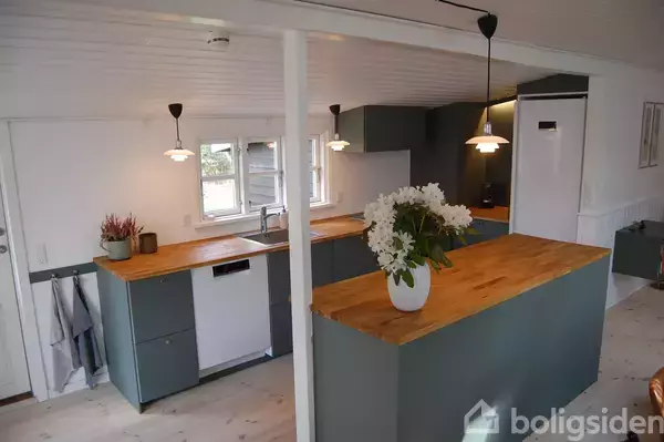 Et køkken med gråt inventar og trælignende bordplader. En vase med hvide blomster står på øen. Loftslamper hænger ned, mens dagslys kommer ind af vinduerne.