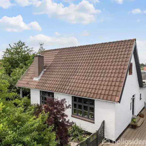 Et hvidt hus med brunt tegltag står blandt grønne træer og buske. Indkørsel med brosten er synlig til højre, omgivet af velplejet have og blå himmel.