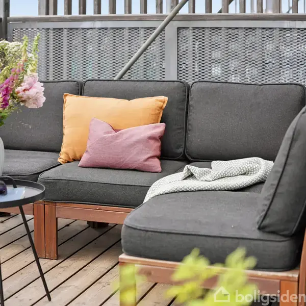 En sort hjørnesofa med farverige puder står på en træterrasse. En lille sort bord viser en vase med blomster og solbriller. Terrasseomgivelser med metalrækværk.