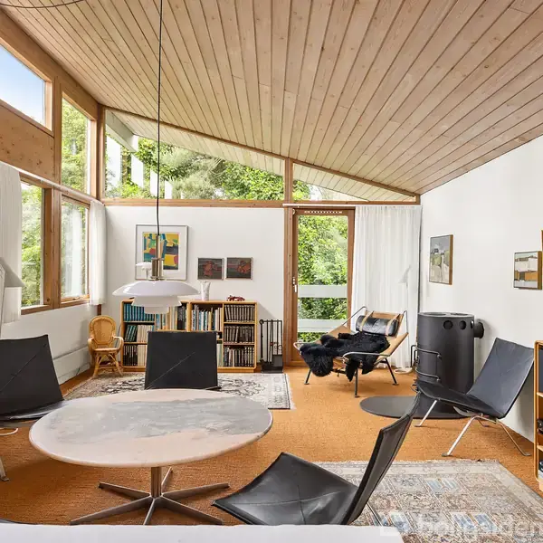 Et moderne opholdsområde med runde stole rundt om et marmorbord. Store vinduer lader naturligt lys ind. Væggene har kunstværker, og reoler er fyldt med bøger.