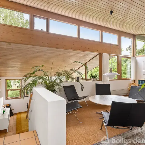 Et moderne, lyst opholdsrum med sorte stole omkring et rundt bord. Store vinduer og træloft tilføjer naturligt lys, mens grønne planter og reoler skaber en hyggelig atmosfære.