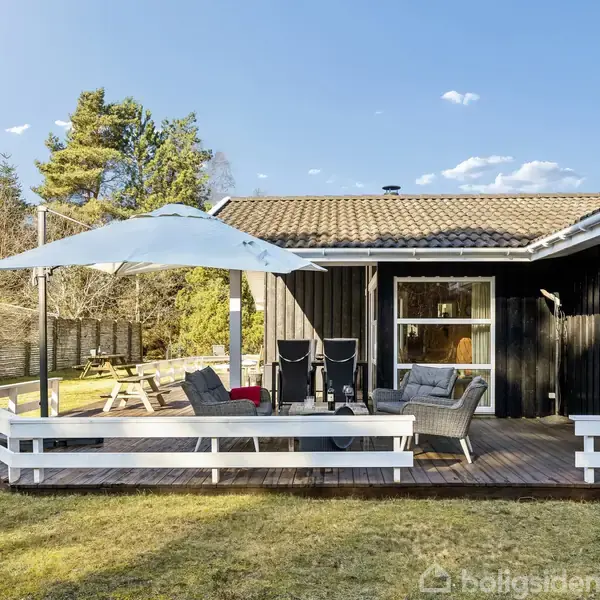 Træterrasse med sofa og parasol, væk fra sortmalet hus. Området er roligt omgivet af buske og træer, med lysende himmel og enkelte skyer.