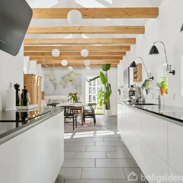 Køkken med hvide skabe og sorte bordplader. Spisebord med stole og planter i baggrunden. Loftet har synlige træbjælker. Verdenskort på væggen og lysindfald fra vinduer.