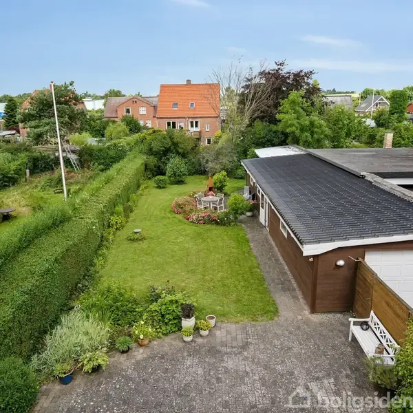 Et parcelhus set fra oven med en grusbelagt indkørsel, garage, velplejet græsplæne og levende hegn. Området er beplantet med buske og træer, omgivet af nabolagets huse.