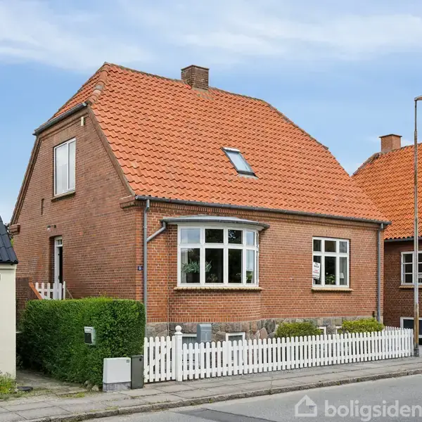 Et rødt murstenshus med rødt tegltag og hvidt stakit står stille ved en vej. Husets store vinduer og skorsten ses klart i et nabolag med lignende huse.