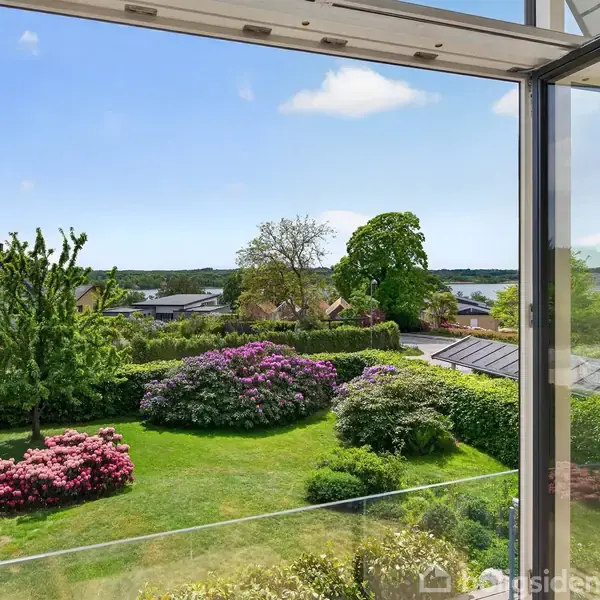 Vindue åbner ud til en have med blomstrende buske og træer. I baggrunden ses et kvarter med huse og en sø under en klar blå himmel.