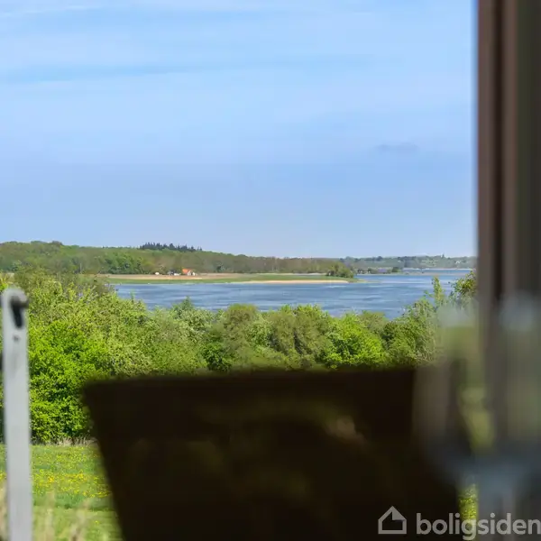 Floden ses gennem et vindue med et glasbord i forgrunden. I baggrunden er der grøn beplantning og en blå himmel, hvilket skaber en fredelig atmosfære.