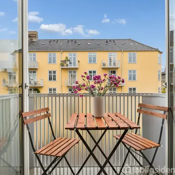 Et lille altanbord med to stole har en vase med lyserøde blomster. Udsigt til gule bygninger med blå himmel i baggrunden.
