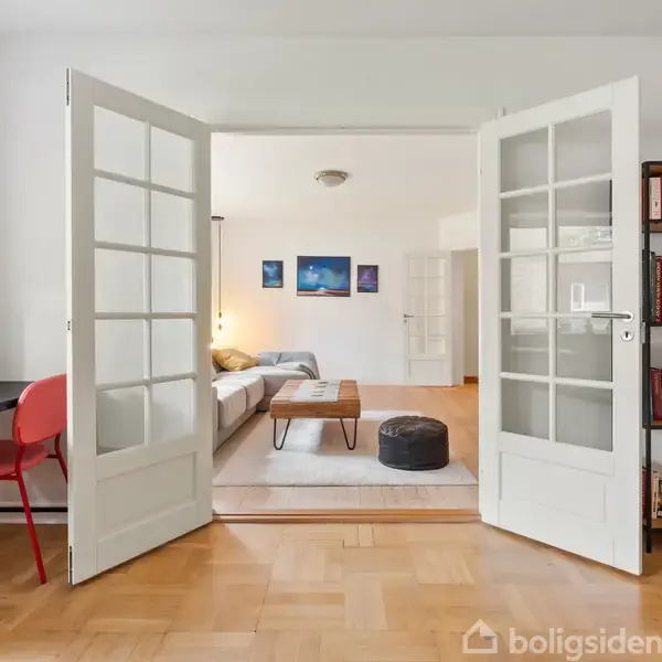 Dobbelt glasdør åbner ind til en stue med en sofa og et sofabord. Væghængte billeder og en bogreol giver en hyggelig atmosfære. En printer står på et bord ved siden af en rød stol.