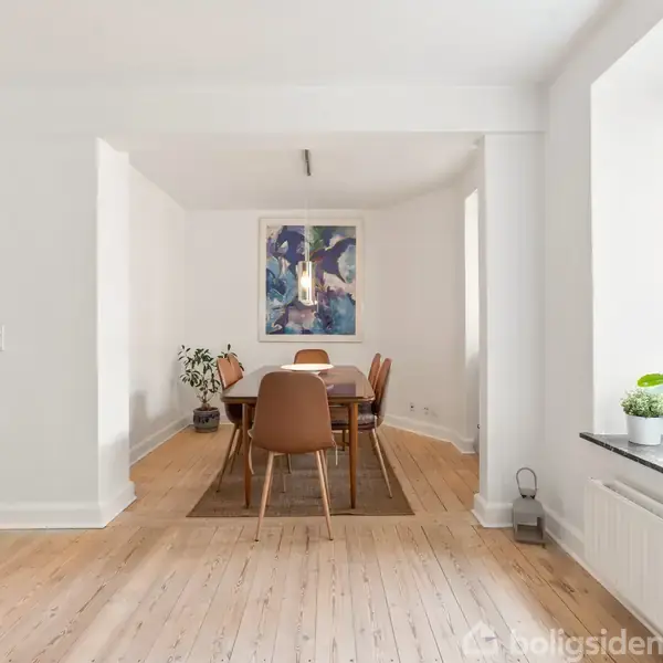 Spisebord med seks stole står på tæppe i rummelig, lys stue. En stor plante og maleri hænger på væggen. Vindue med udsigt til bygninger og planter på vindueskarmen.