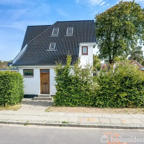 Hvidt hus med sort tag, stående bag en hæk. Huset har flere vinduer og en trædør. Det ligger i et villakvarter med fortov, træer og vejskilte.