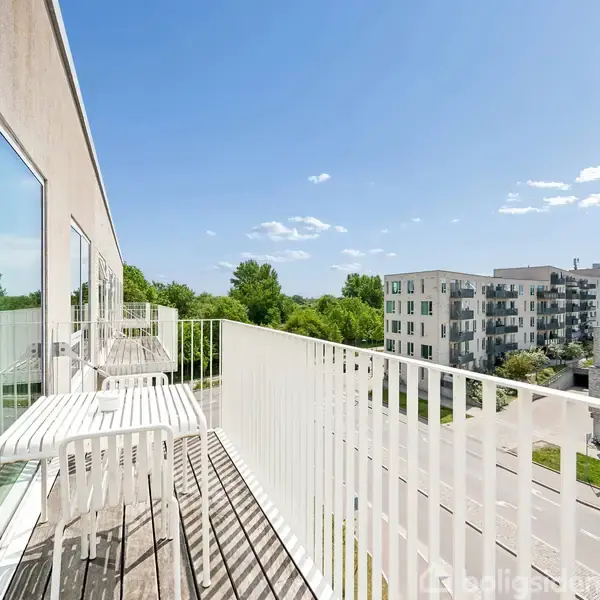 En balkon med et hvidt bord og to stole står mod en glasvæg. Den har udsigt over moderne bygninger og grønne træer under en blå himmel.