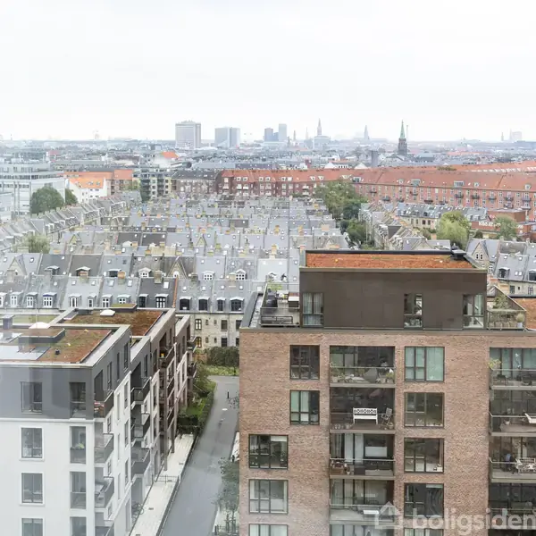 Højhus set udefra, roligt uden aktivitet, omgivet af andre tætpakkede lejlighedsbygninger og hustage i en urban byscene. Vandtårne og kirketårne skimtes i horisonten.