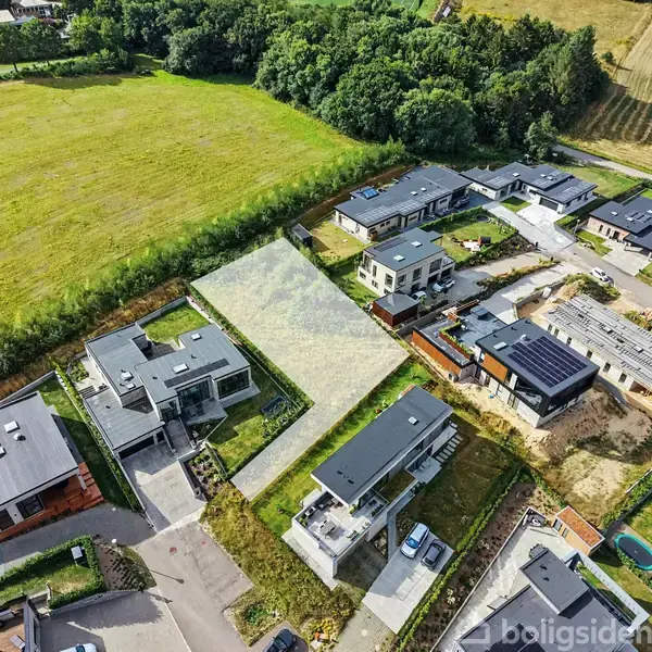 Boligkvarter med moderne huse set fra luften. Husene har flade tage og er arrangede langs en vej. Omkringliggende marker og skov danner en grøn baggrund.