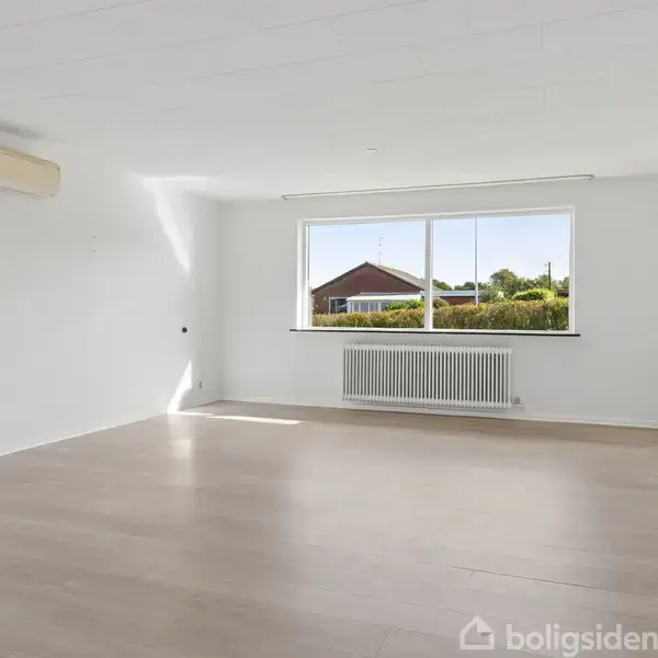 Tomt rum med en stor vindue, hvide vægge og lyse trægulve. Vindue viser en udsigt over et hus og grønne omgivelser udenfor. Radiator placeret under vinduet.