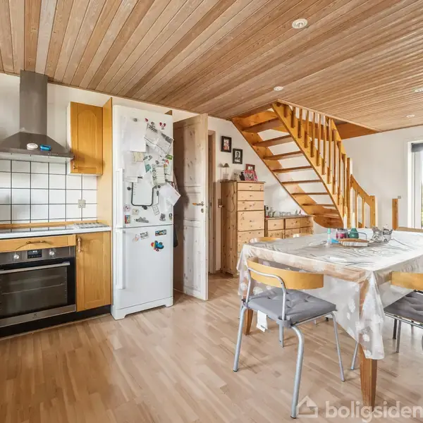 Køkken med spisebord og stole; køleskab med magneter; trappe fører opad; træloft og parketgulv; store vinduer giver udsigt til have.
