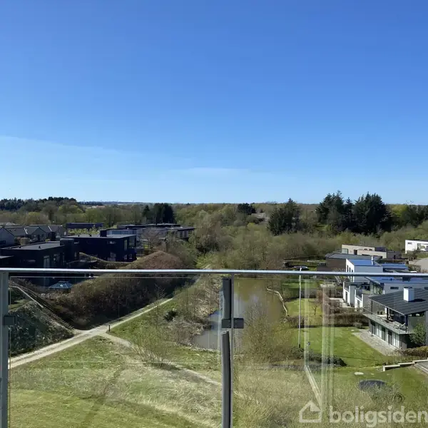 En glasbalkon med udsigt over et boligområde omgivet af grønt landskab og skovtræer under en klar blå himmel.