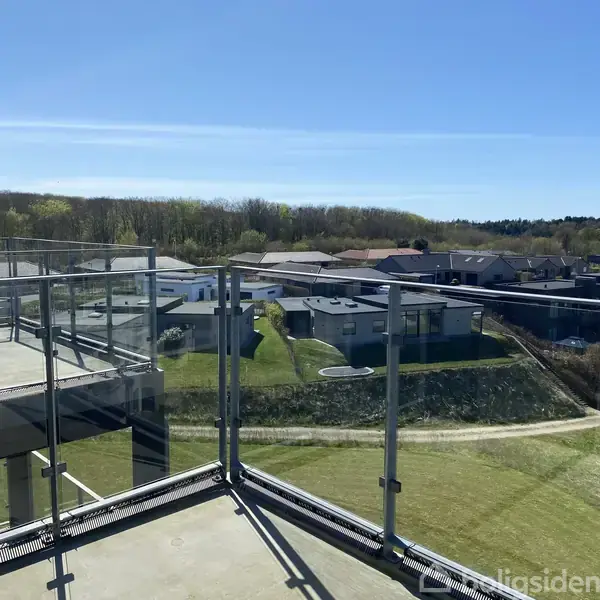 En altan med glasrækværk står stille og tilbyder udsigt over et boligområde med moderne huse og grønne omgivelser under en klar, blå himmel.