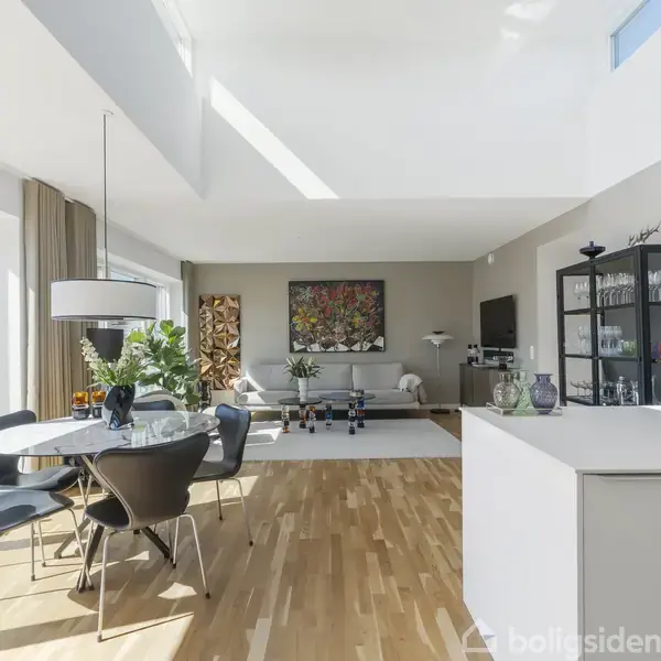 Moderne stue med spisebord og stole står ved siden af et stort vindue, der lader sollys skinne ind. Lyse trægulve, stilfulde møbler og kunstværk på væggen skaber en elegant atmosfære.
