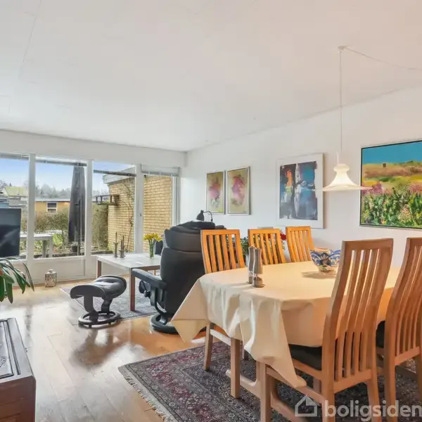 Et spisebord med otte stole står i en lys stue. Stuen har store vinduer, en sofa, en lænestol og farverige vægmalerier. et tæppe ligger under bordet.