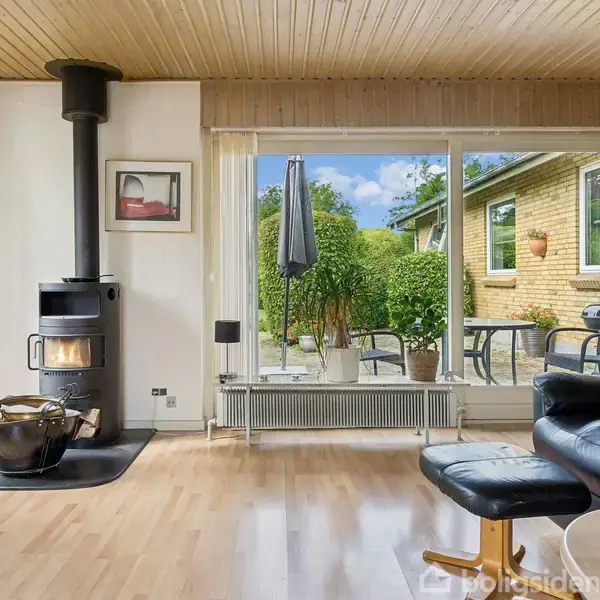 En brændeovn står til højre og opvarmer stuen med trægulv. En stor glasdør med udsigt til en terrasse og have udgør baggrunden. Sort lænestol i forgrunden.