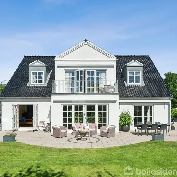 Hus med hvid facade og sort tag. På terrassen står havemøbler, og en swimmingpool ses til højre. Græsplæne og træer i baggrunden under en klar blå himmel.