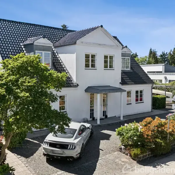 Hvidt hus med sort tegltag står i et boligkvarter omgivet af grønne træer og buske. En grå bil er parkeret på grusindkørslen foran huset.