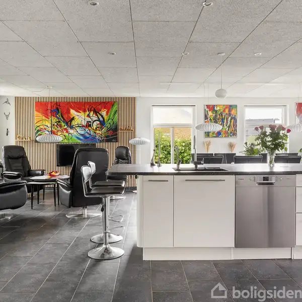 Køkken-alrum med moderne indretning: Hvide skabe, grå flisegulv, og en stor køkkenø med barstole. Lyse vægge prydet med farverige malerier. Store vinduer giver naturligt lys.