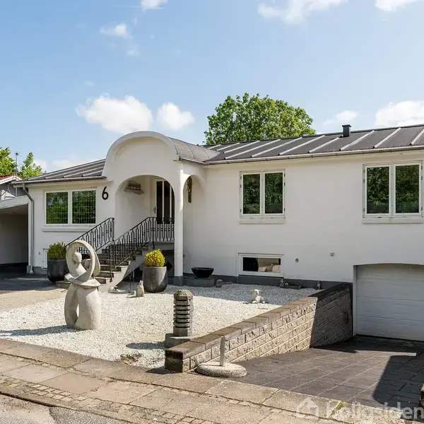 Hvidt hus med moderne skulptur ved indgangen, beliggende i et beboelsesområde med garage og grusgård. Husnummer 6 vises tydeligt ved indgangen.