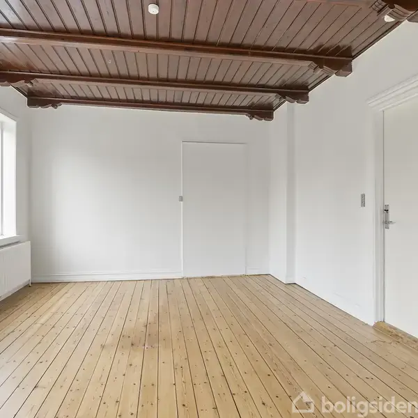 Et tomt rum med trægulv og hvidmalede vægge. Mørke træbjælker pryder loftet. Til venstre er der et vindue med udsigt og en radiator.