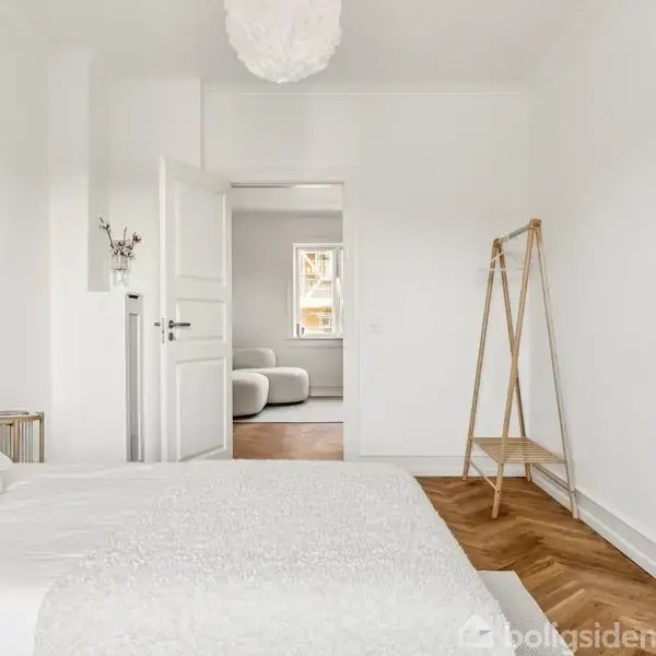 Et soveværelse domineret af en hvid seng står i fokus, med en åben dør der fører til en lys stue. Væggene er hvide, og der er et trælaminat gulv.