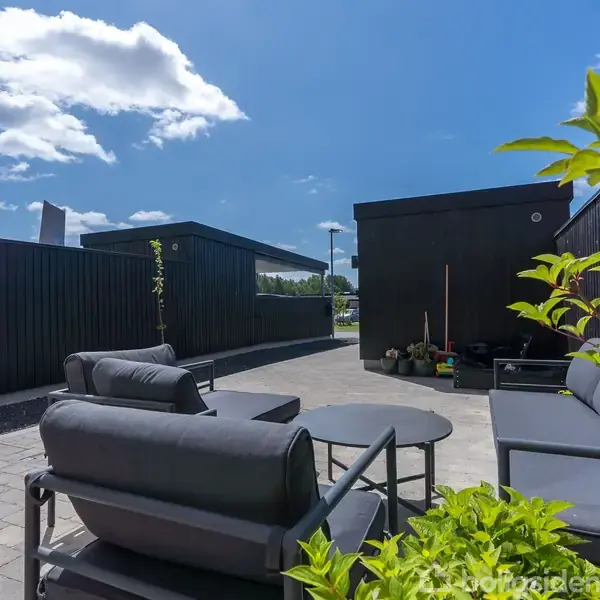 Sorte udendørs lounge-møbler står på en flisebelagt terrasse omgivet af mørke træpaneler under en blå himmel med skyer. Grønne planter tilføjer et strejf af natur.