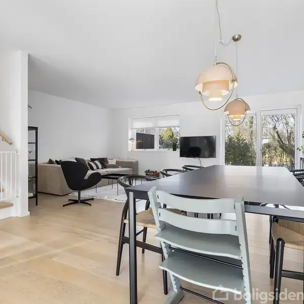 En lys spisestue med et sort bord omgivet af stole, overhængt af pendellamper. Ved siden af er en stue med sofaer, sofabord og fladskærms-TV. Store vinduer viser grønne træer udenfor.