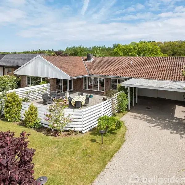 Hus med rødt tag og hvidt træhegn har en terrasse med havemøbler. Foran huset ses en carport og en stor have med forskellige buske og træer.
