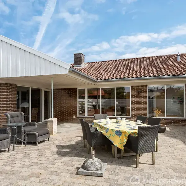 Terrasse med spisebord dækket med en gul dug og seks stole under åben himmel. Omgivet af murstenshus med store vinduer og udendørs siddeområde med to stole.